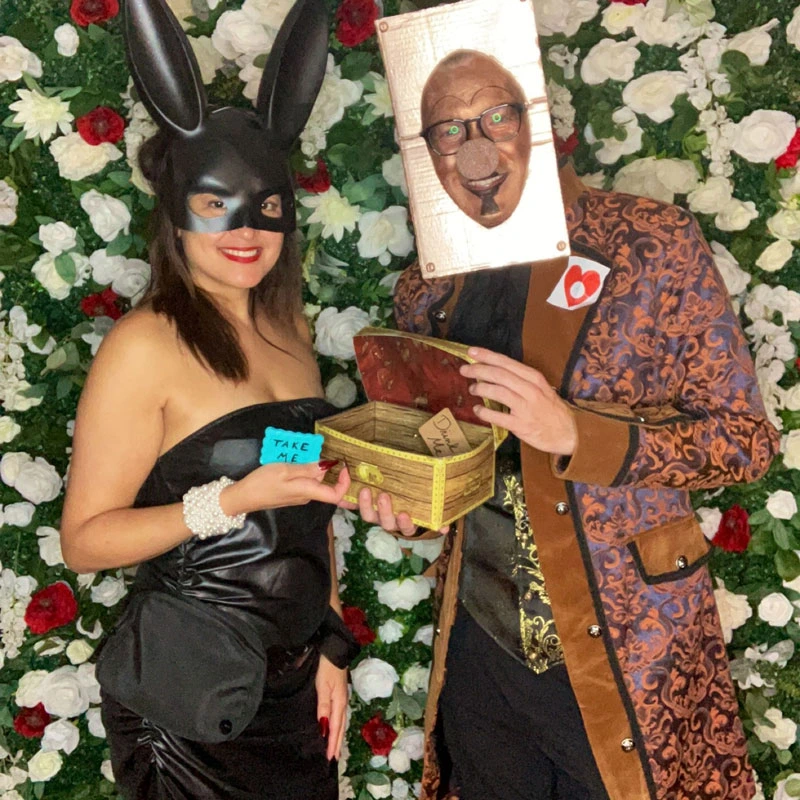 Man and woman couple, she's dressed as a rabbit with a black mask, he is dressed in a mask that makes his face look like an ornate doorknob; they're holding a box with cards that say 'drink me'